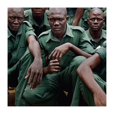 A group of African men in green uniforms, likely soldiers, are closely gathered, gazing directly forward with serious expressions in an outdoor setting.