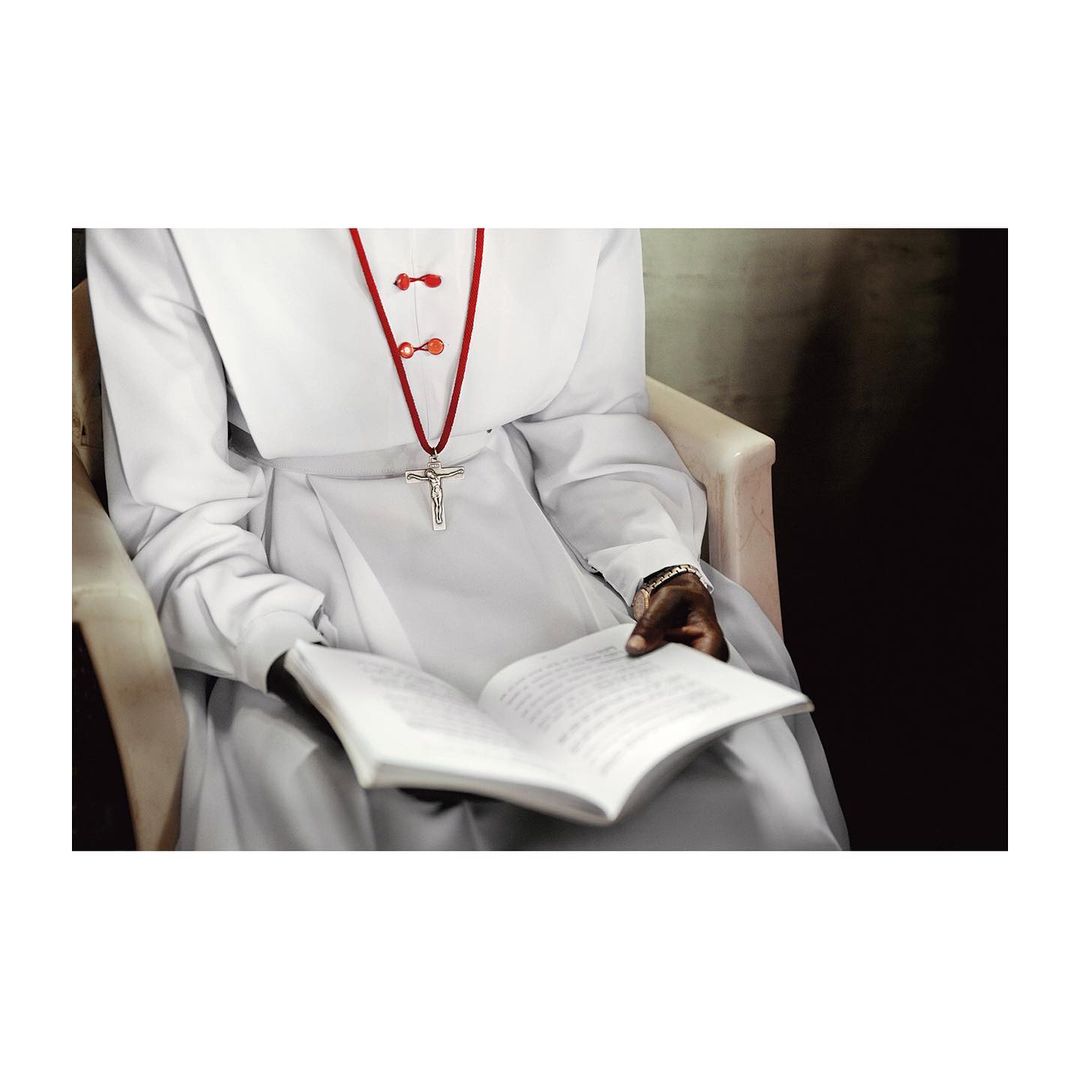 Nun in white habit seated and reading a book with a crucifix pendant on a red cord; dark background with chair visible.