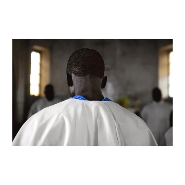 An adult male in white religious clothing stands with his back to the camera, in a room with other adults and natural light.