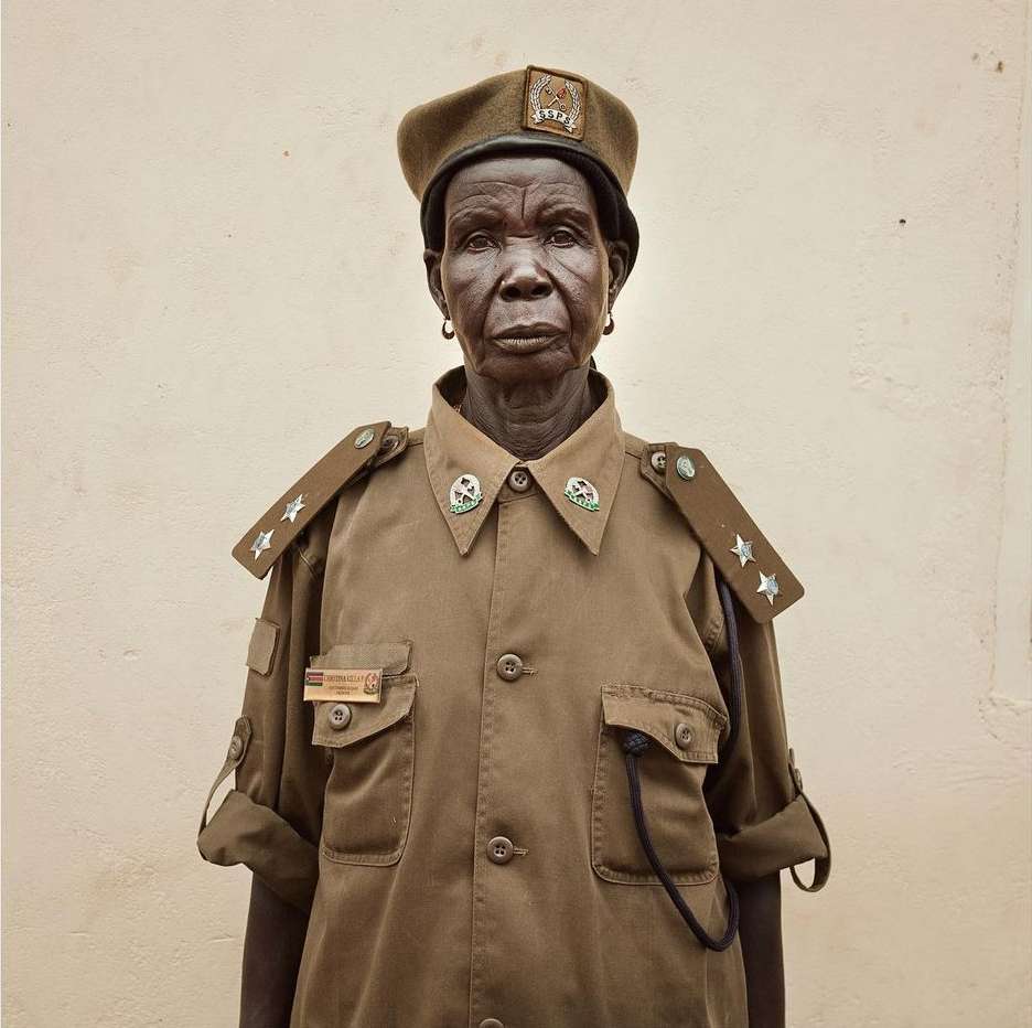 Close-up of an elderly African woman in a tan uniform and beret, featuring a neutral facial expression against a plain light-colored background.
