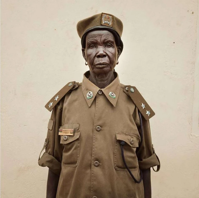 Close-up of an elderly African woman in a tan uniform and beret, featuring a neutral facial expression against a plain light-colored background.
