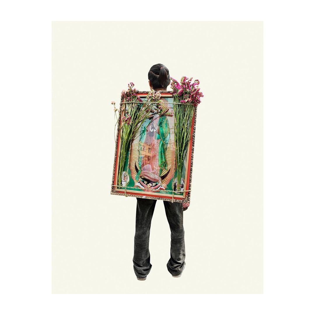 Person carrying a framed painting of the Virgin Mary adorned with flowers on their back in a studio shot against a cream background.