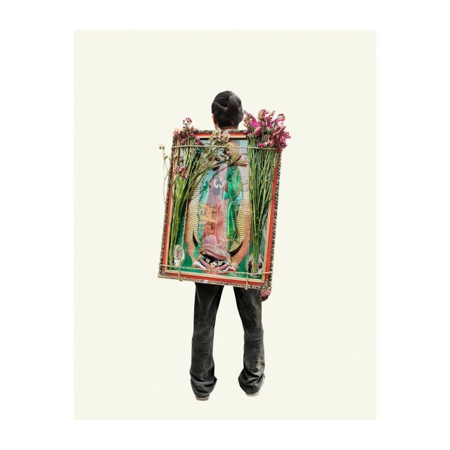 Person carrying a framed painting of the Virgin Mary adorned with flowers on their back in a studio shot against a cream background.
