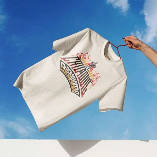 T-shirt with printed design on a hanger against a blue sky with clouds, featuring visible arm holding hanger.
