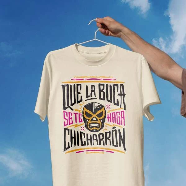 T-shirt featuring luchador mask graphic and Spanish text, held up against a bright blue sky with wispy white clouds creating backdrop.