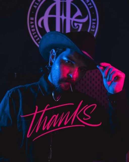 Bearded man in cowboy hat wearing a black shirt with 'Thanks' printed on it, neon-lit face, and a decorative background emblem.
