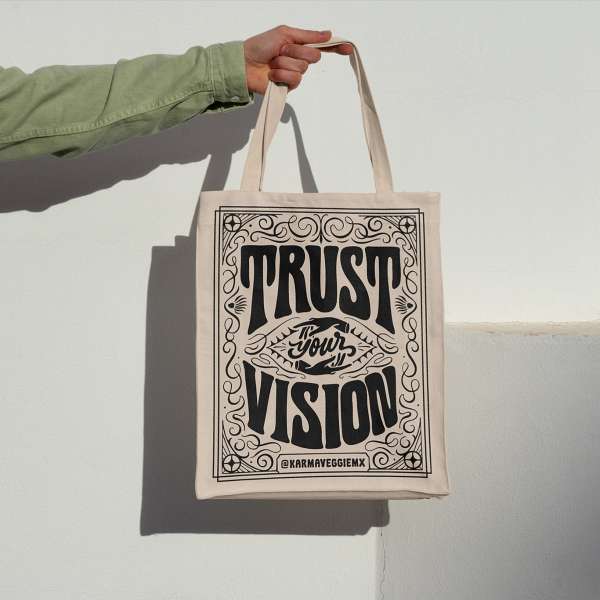 Hand holding a beige tote bag with black lettering that says 'Trust your vision' in an indoor setting against a white wall.