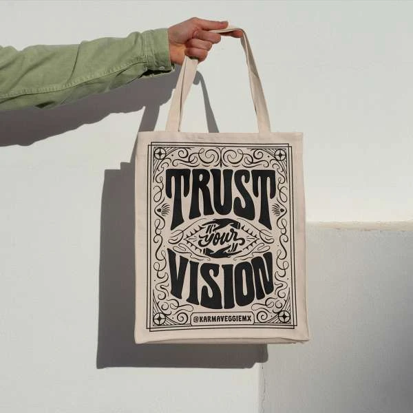 Hand holding a beige tote bag with black lettering that says 'Trust your vision' in an indoor setting against a white wall.