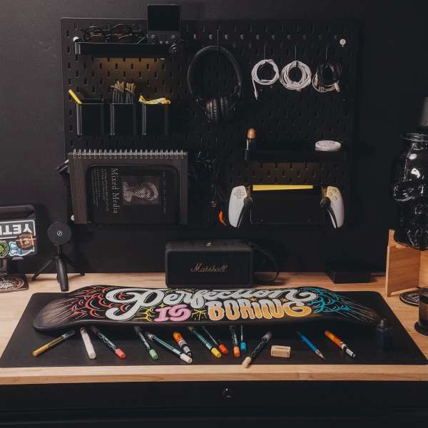 Skateboard art on desk with markers and black pegboard background holding headphones, controller, cables, and a Marshall speaker.
