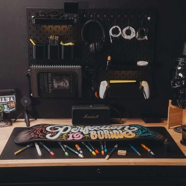Skateboard art on desk with markers and black pegboard background holding headphones, controller, cables, and a Marshall speaker.