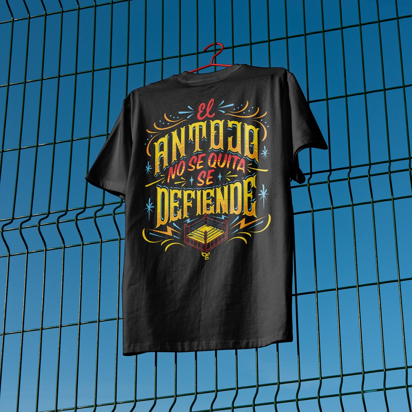 Black t-shirt with 'El Antojo No Se Quita Se Defiende' text and boxing ring graphic on a hanger in front of a blue fence.