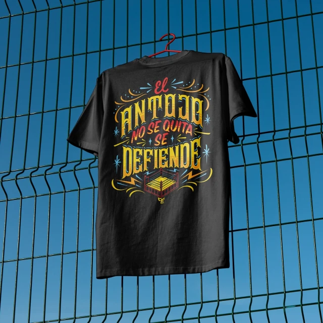Black t-shirt with 'El Antojo No Se Quita Se Defiende' text and boxing ring graphic on a hanger in front of a blue fence.