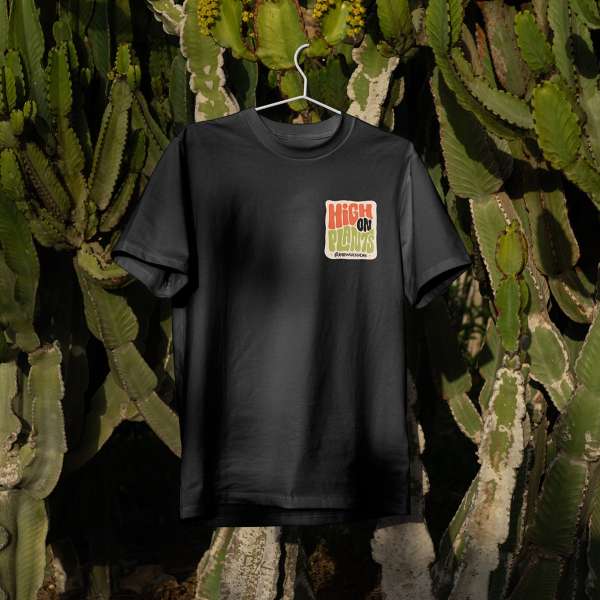 Black t-shirt on a hanger with a graphic patch saying 'High On Plants', displayed against a backdrop of green cactus plants.