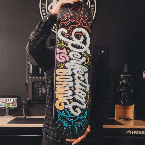 Person holding skateboard with the text 'Perfection is Boring'. Black background with desk, pegboard, Yeti cooler and decorative items.