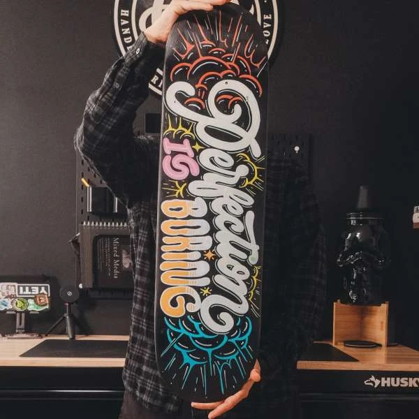 Person holding skateboard with the text 'Perfection is Boring'. Black background with desk, pegboard, Yeti cooler and decorative items.
