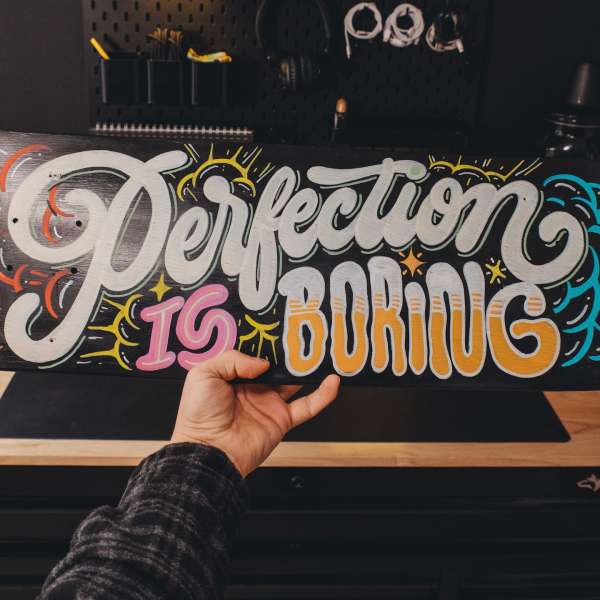 Close-up of a hand holding a skateboard deck with the hand-painted typography, 'Perfection is Boring' on a dark background.