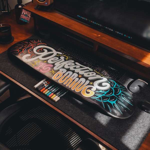 Artistic skateboard featuring the phrase 'Perfection is Boring' in colourful hand-lettering, markers and a monitor in the background.