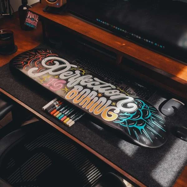 Artistic skateboard featuring the phrase 'Perfection is Boring' in colourful hand-lettering, markers and a monitor in the background.