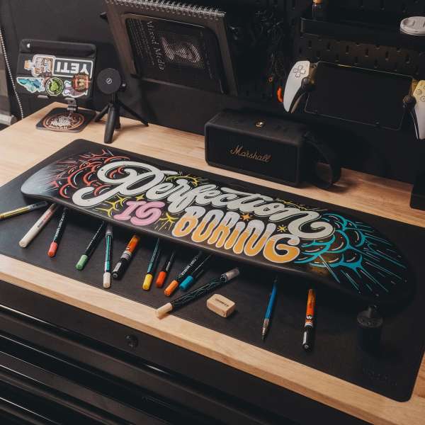 A skateboard with painted typography and colorful details sits among markers, a speaker, and digital devices in a tidy indoor setting.