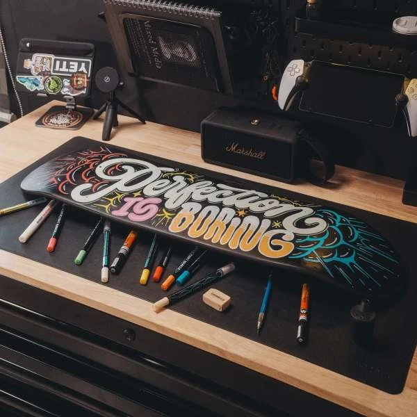 A skateboard with painted typography and colorful details sits among markers, a speaker, and digital devices in a tidy indoor setting.