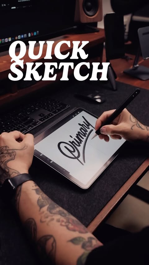 Hand with tattoos uses a stylus on a tablet to draw the word 'Primary'. Keyboard, speaker, and desk are visible in soft studio light.