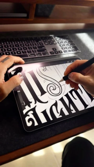 Close-up of hands using a stylus on a tablet to create digital art with bold lettering, indoors on a wooden desk with a keyboard.