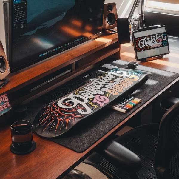 Desk setup with a skateboard, computer monitor, speakers, tablet, and markers on a wooden desk with a dark gray mat.