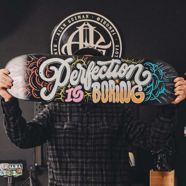 Person in plaid shirt holding skateboard with 'Perfection is Boring' graphic art in front of face, indoors with logo in background.