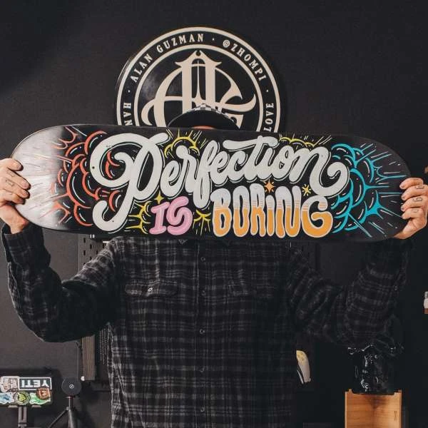 Person in plaid shirt holding skateboard with 'Perfection is Boring' graphic art in front of face, indoors with logo in background.