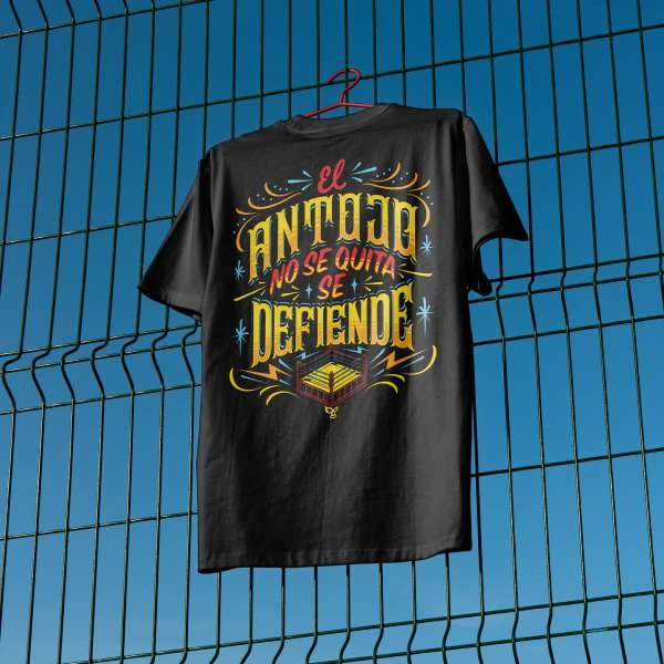 Black t-shirt on a hanger against a blue sky and green fence features Spanish text and graphic of a wrestling ring.