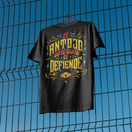 Black t-shirt on a hanger against a blue sky and green fence features Spanish text and graphic of a wrestling ring.