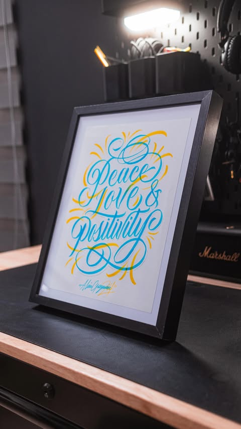 Framed wall art on a wooden desk featuring a handwritten-style typography design in teal and yellow with the message Peace, Love, & Positivity.