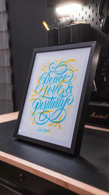 Framed wall art on a wooden desk featuring a handwritten-style typography design in teal and yellow with the message Peace, Love, & Positivity.