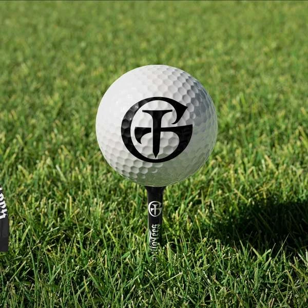 Close-up of a white golf ball with a black logo on a black tee, set in short, green grass. Focused sports equipment scene.