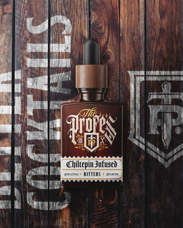 Overhead studio shot of a dark brown bitters bottle with a wood grain label, against a wooden background with white painted text.