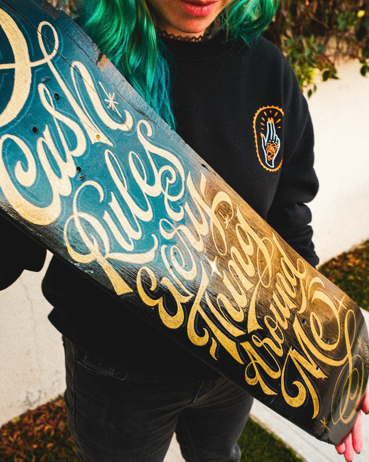 Skateboard art featuring hand-painted lettering in blue and gold hues, held by a woman with teal hair in a casual outdoor setting.