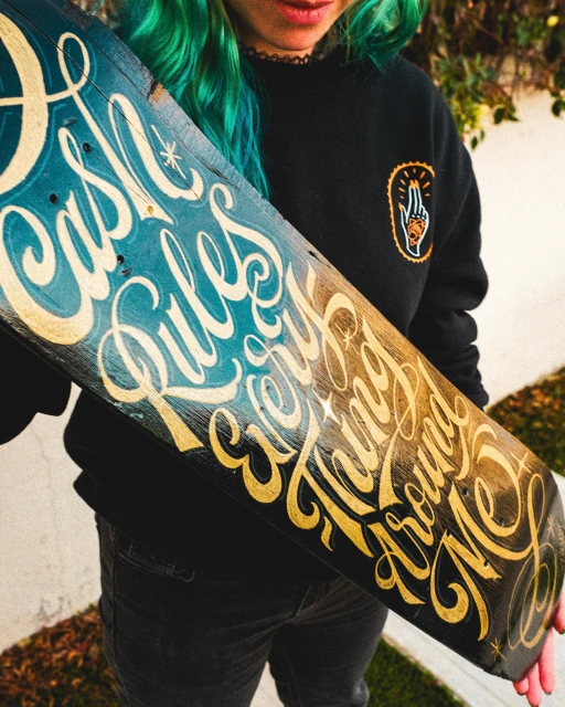 Skateboard art featuring hand-painted lettering in blue and gold hues, held by a woman with teal hair in a casual outdoor setting.