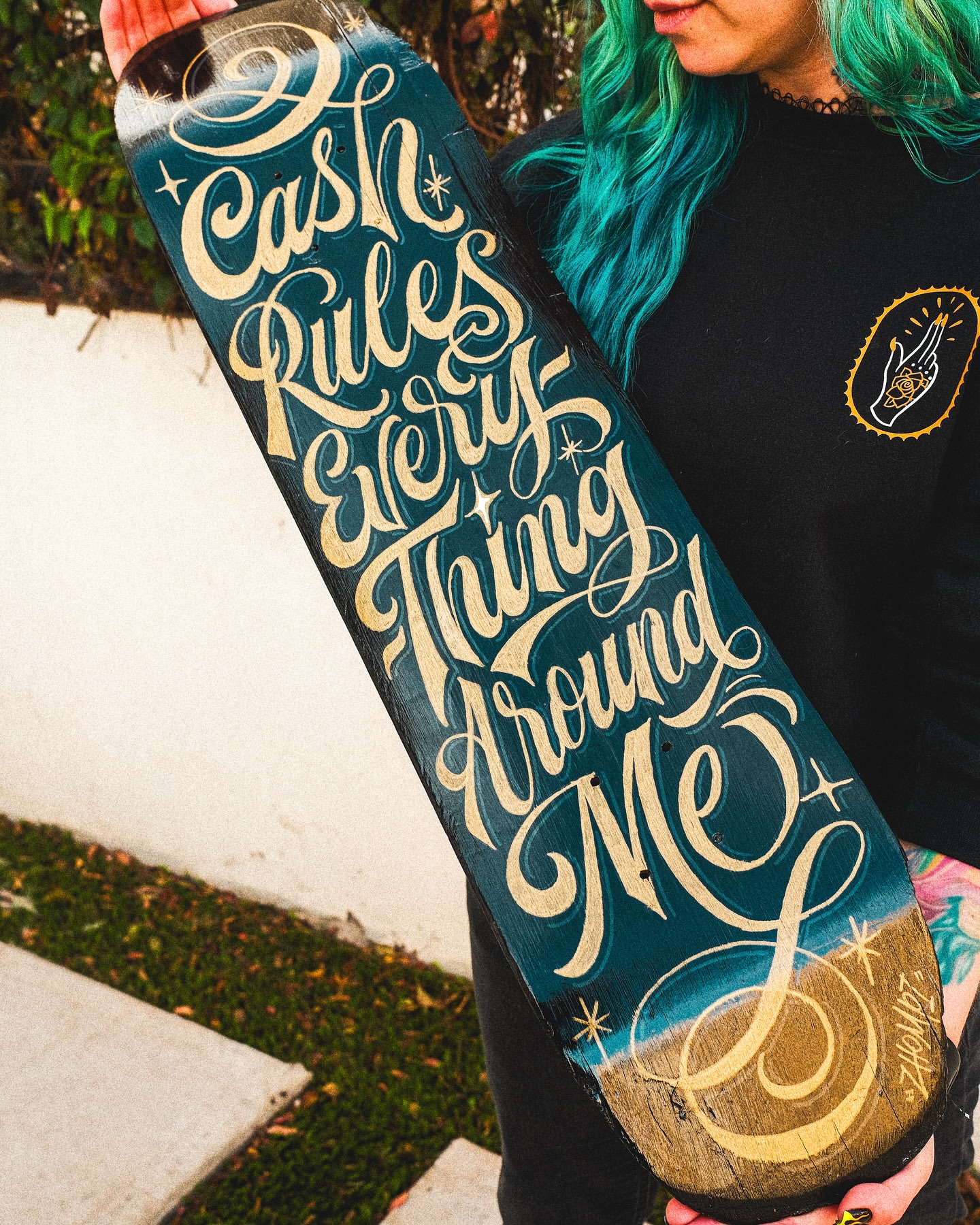 Close-up of a person holding a skateboard with turquoise and gold text that reads, Cash Rules Everything Around Me. 