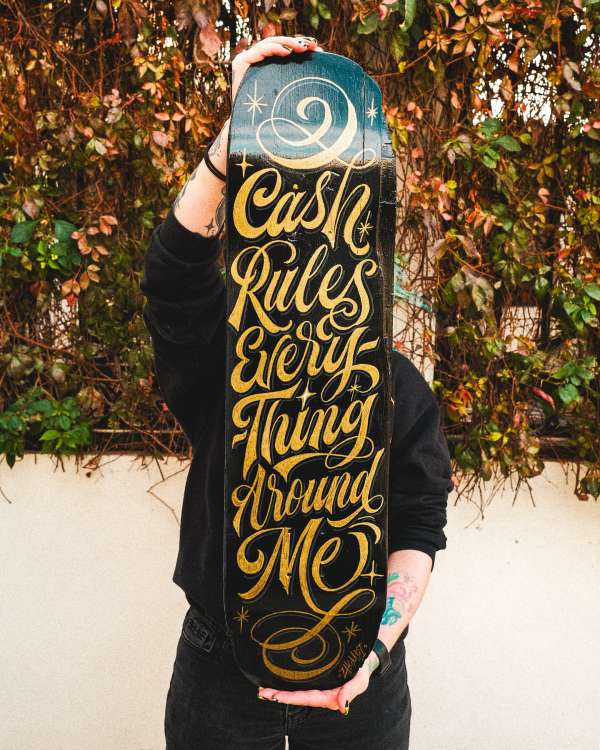 Person holding a skateboard with 'Cash Rules Everything Around Me' lettering in a close-up shot, outdoor setting with a lush green background.