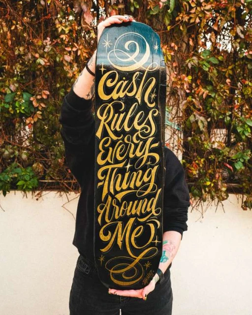 Person holding a skateboard with 'Cash Rules Everything Around Me' lettering in a close-up shot, outdoor setting with a lush green background.