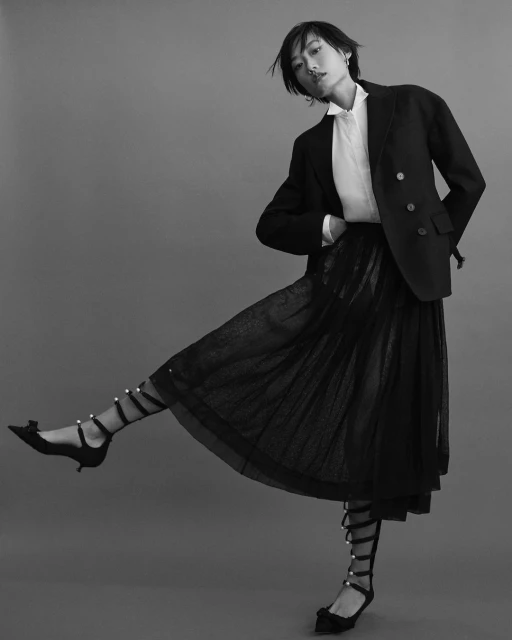 Elegant woman in a black blazer and lace skirt strikes a pose against a grey backdrop, showcasing high-fashion style and poise.