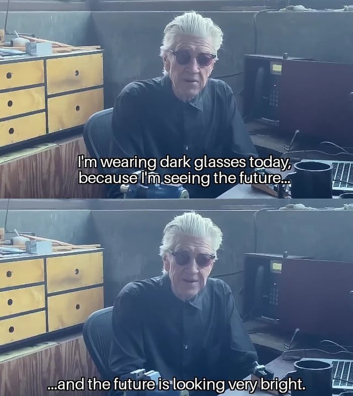 Older man with white hair wearing dark glasses and a black shirt sitting in an office environment looking at the camera.