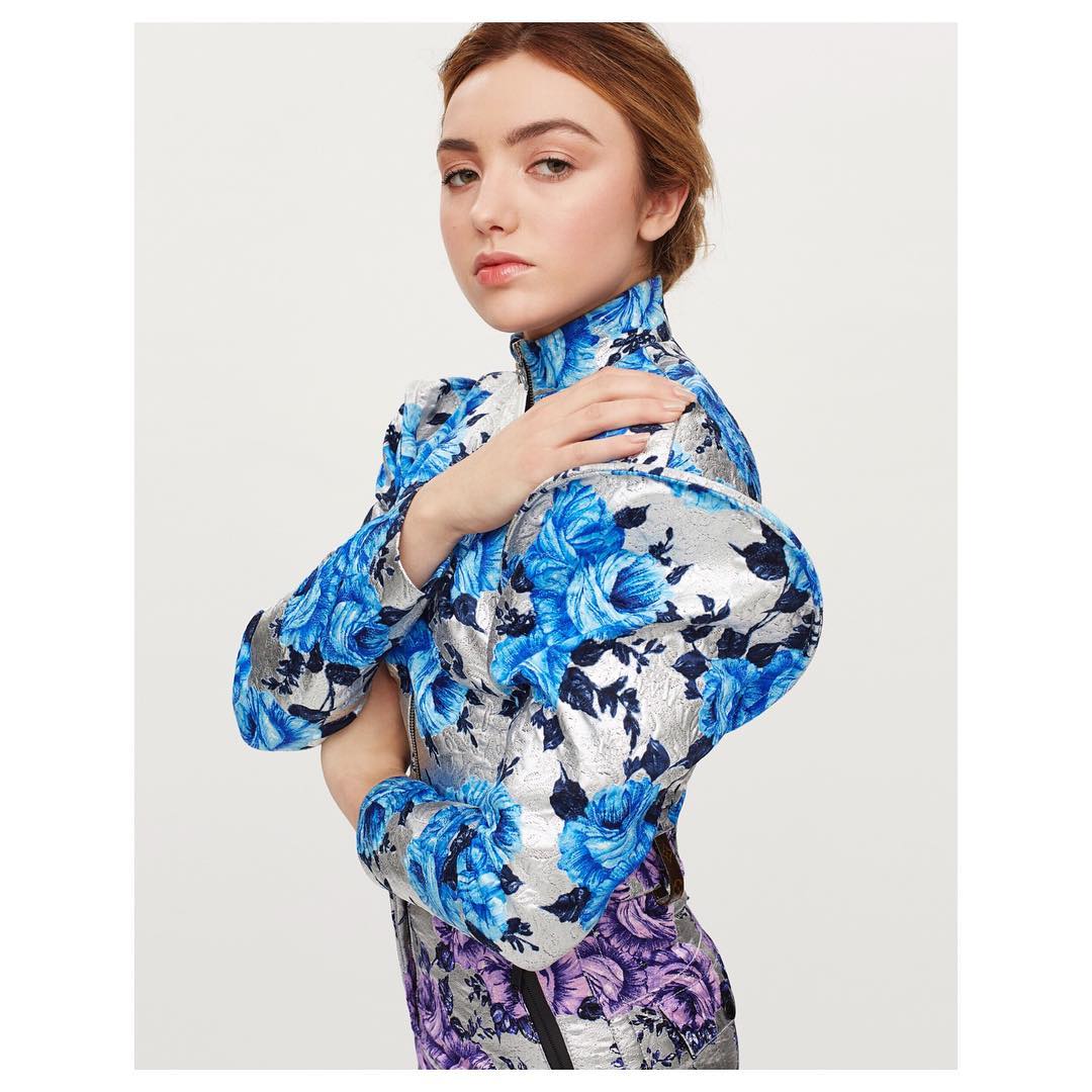 Attractive young woman poses indoors in a floral-patterned dress with puffy sleeves against a plain white backdrop. Fashion photography.
