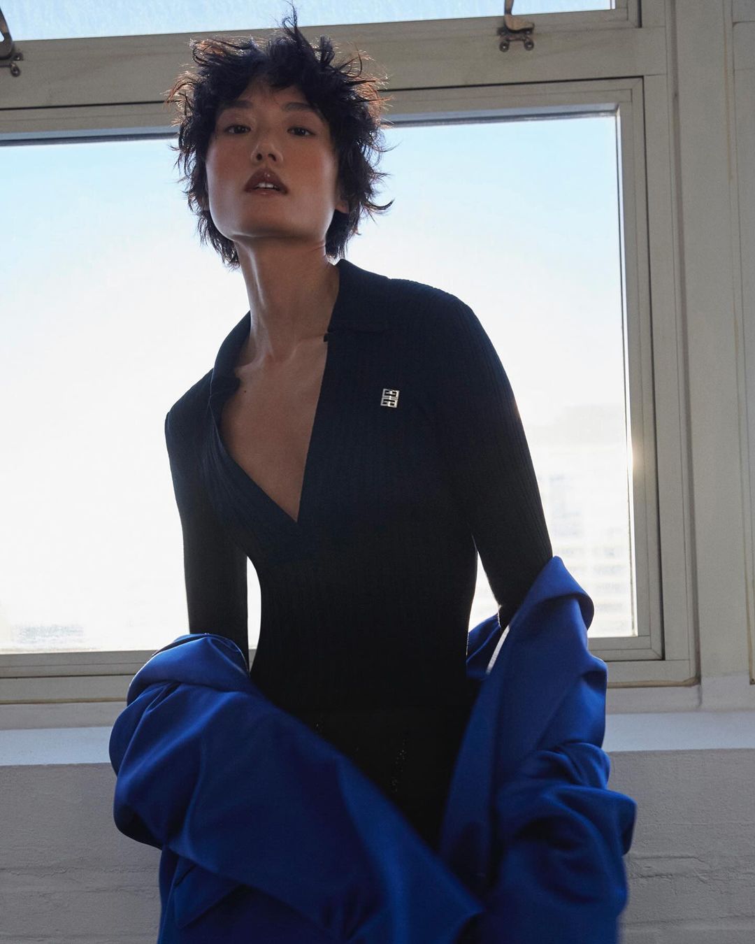 Portrait of an Asian female fashion model in a black top and blue jacket, standing near a window with city views in the background.