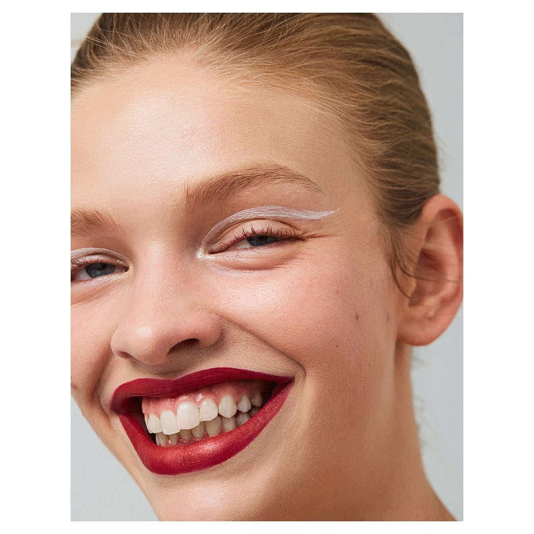 Close-up of a smiling young woman with fair skin, bright red lipstick, white eyeliner, and white teeth against a neutral background.