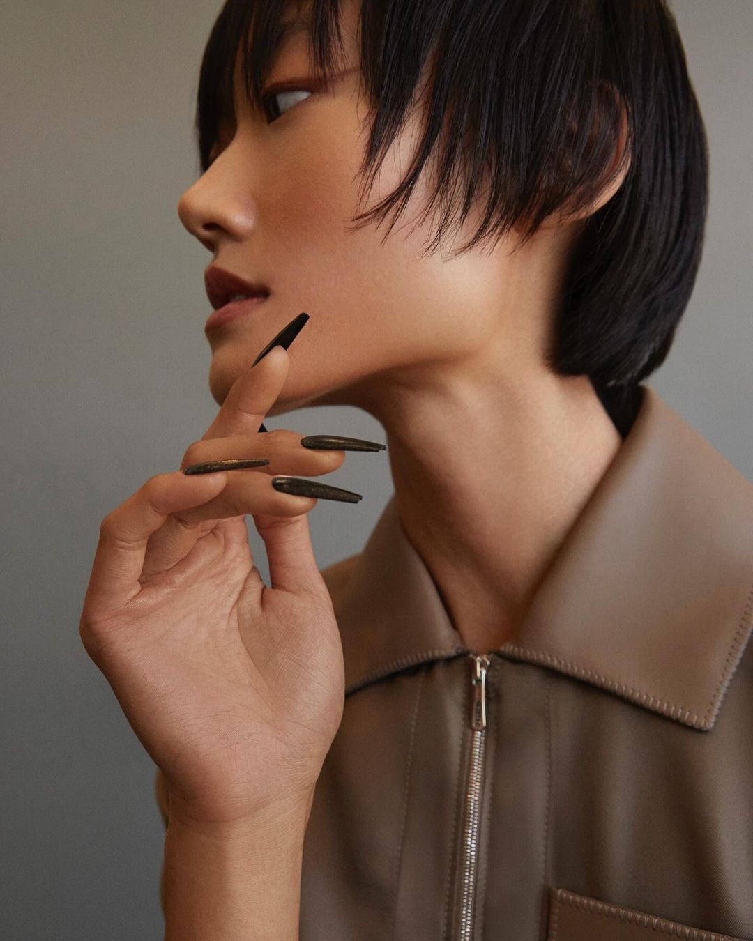Close-up studio shot of a young Asian female model with short black hair, wearing a tan shirt, and posing with her hand near her face.