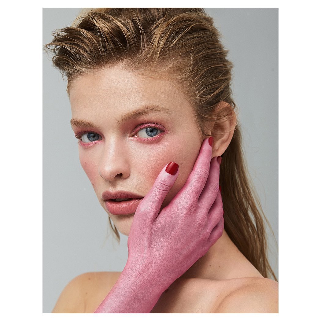 Close-up portrait of a young woman with pink-toned skin and makeup, fair hair, and light eyes in a minimalist studio setting.