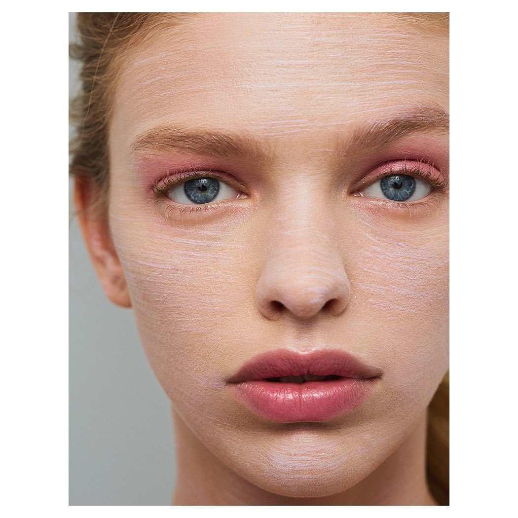 Close-up of a woman's face with a pale cosmetic mask, pink eyeshadow, and light pink lips against a gray background.