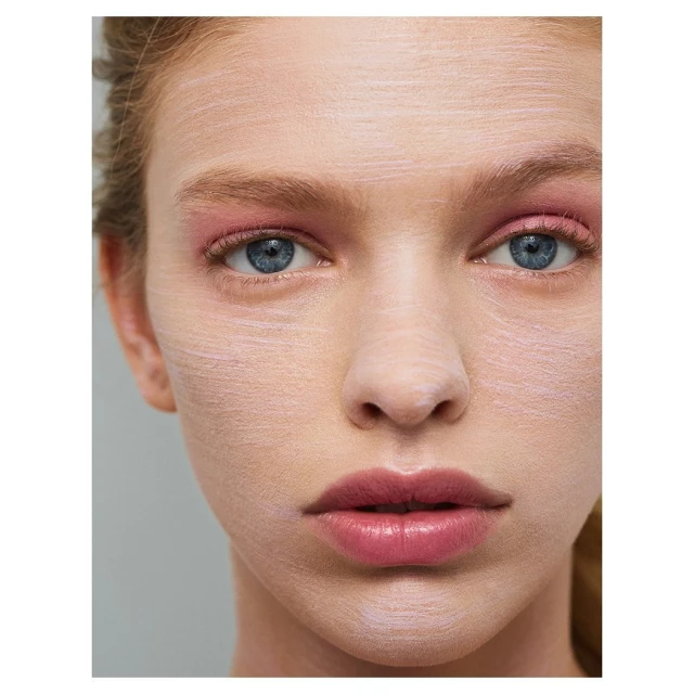 Close-up of a woman's face with a pale cosmetic mask, pink eyeshadow, and light pink lips against a gray background.