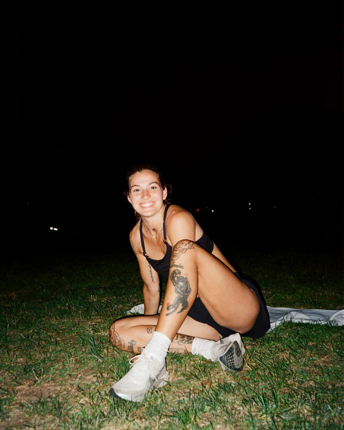 Woman with tattoos sits on grass at night in athletic attire, smiling with white running shoes. Dark sky with subtle city lights.